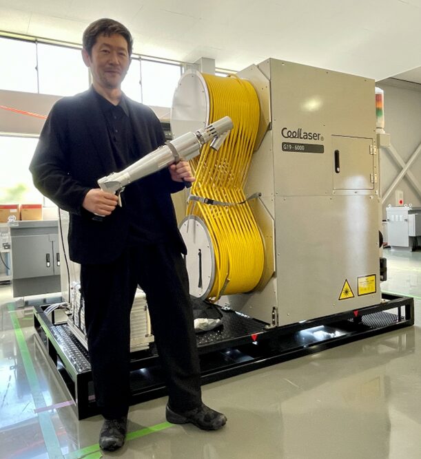 写真１：“出荷を待つ5.4kW出力CoolLaserの前でレーザーヘッドを持つ【株式会社トヨコー】豊澤一晃代表取締役CEO”。2026年4月、CoolLaser製造・メンテナンス拠点HAMAMATSU BASE（〒434-0041静岡県浜松市浜名区平口657-1）にて撮影